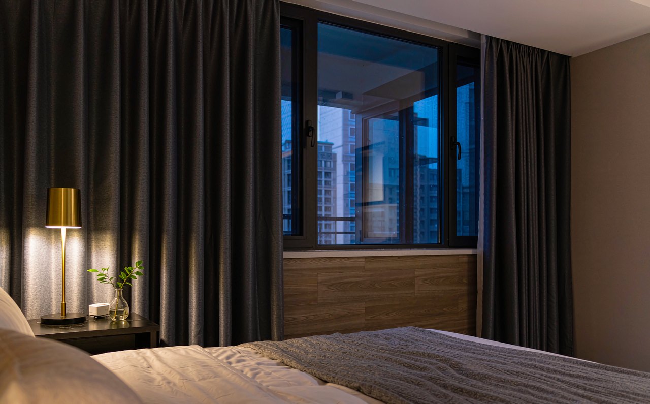 A tidy cozy bedroom at dusk with warm bedside lamp light and blackout curtains