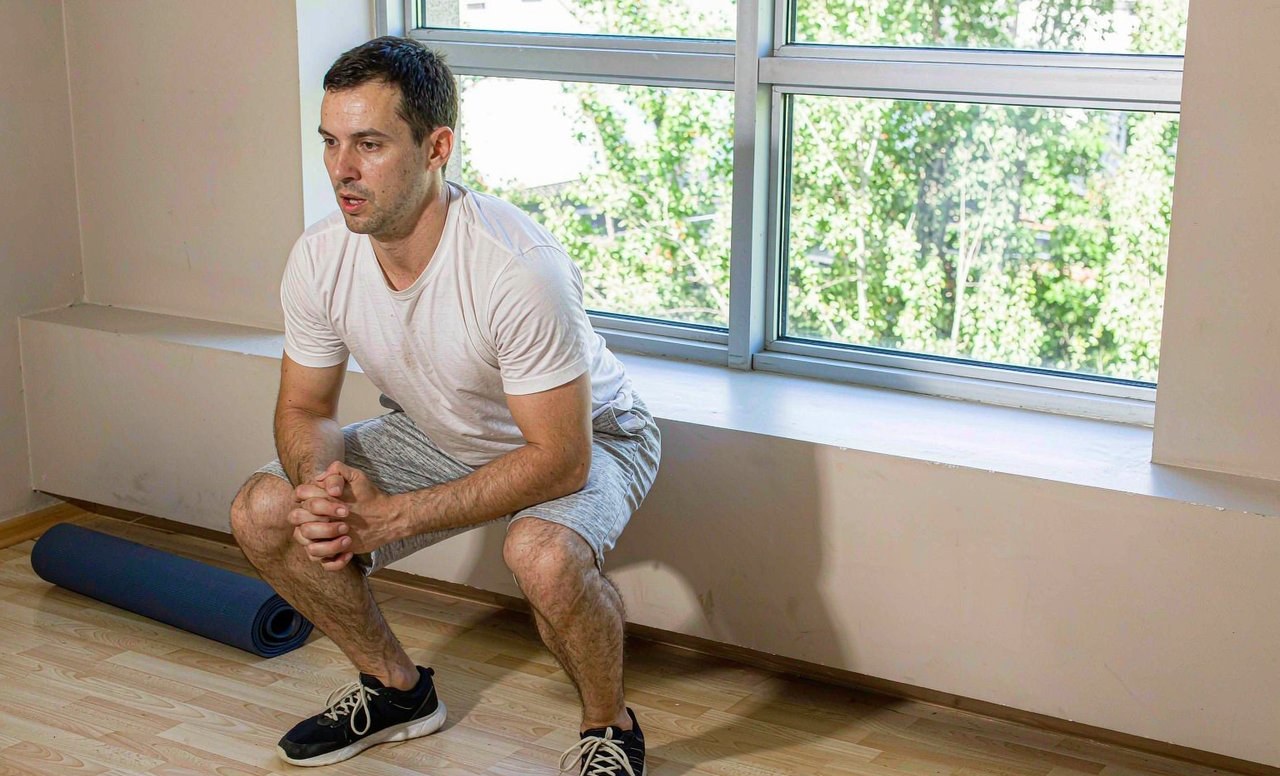 A man performing a bodyweight squat in a bright living room beside a rolled-up exercise mat and a water bottle