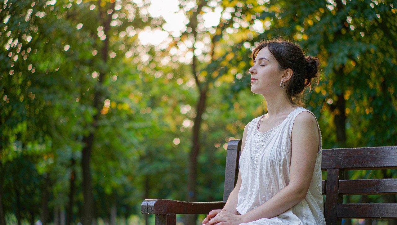 A woman sitting calmly on a park bench with her eyes closed, hands resting on her lap, soft golden afternoon light through green trees, realistic photo style