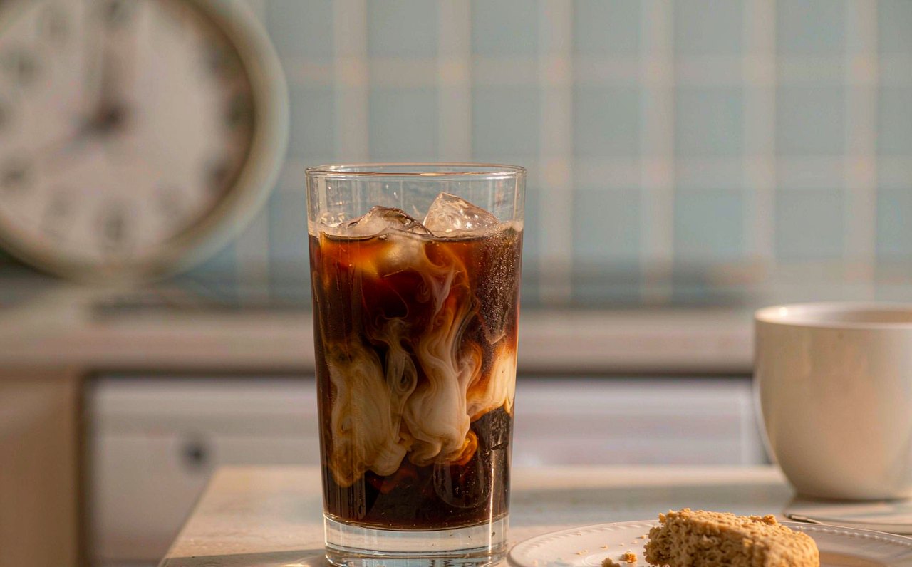 A half-full glass of iced coffee on a kitchen counter beside a wall clock, soft afternoon light across the scene