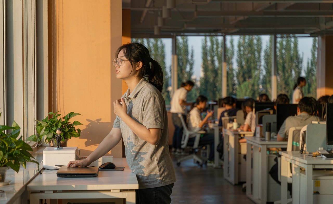 An office worker with glasses standing at her tidy desk, looking out of a large window into the distance during a screen break in warm afternoon light