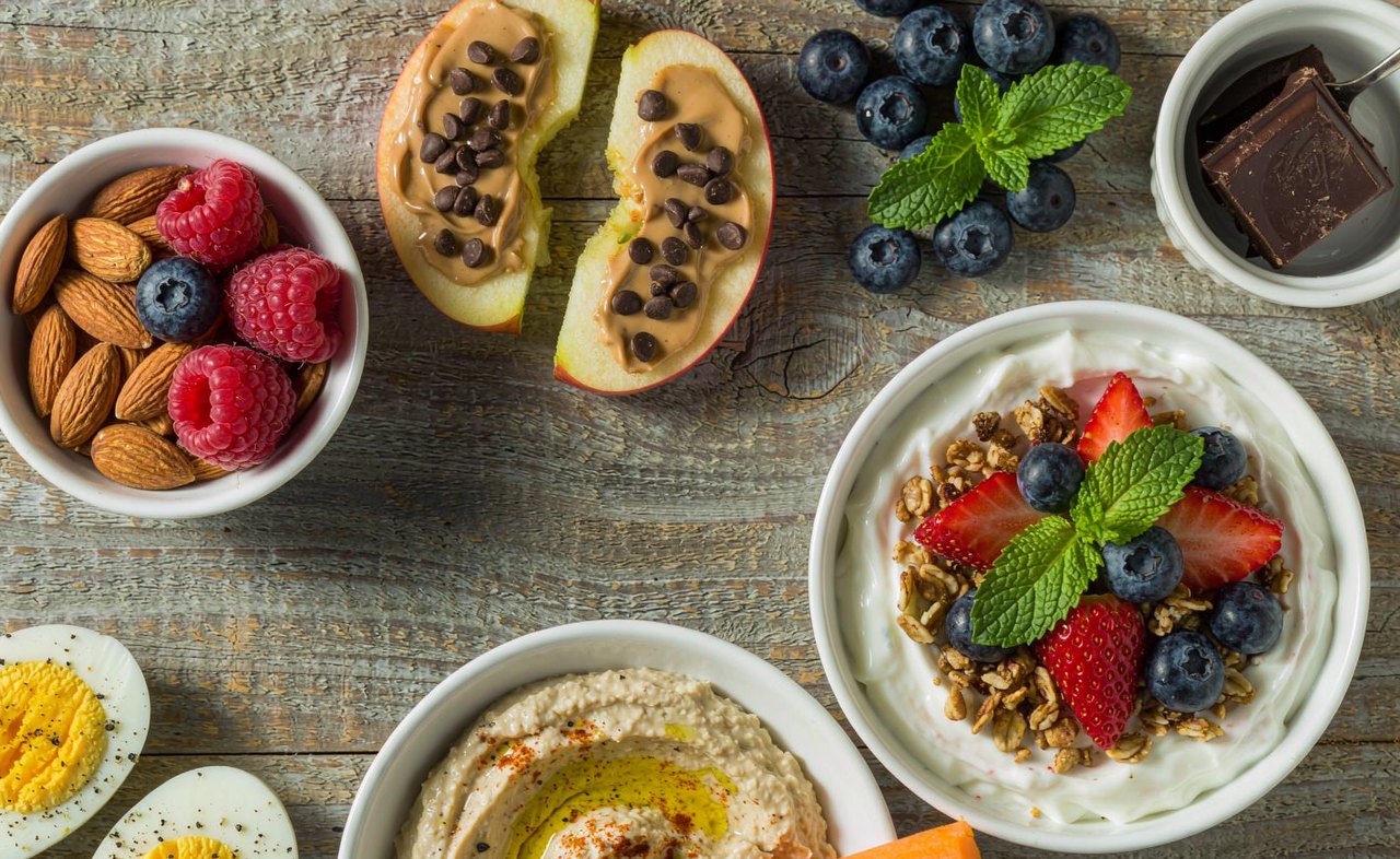 An overhead view of healthy snacks on a wooden table including apple slices, almonds, yogurt and carrot sticks with hummus