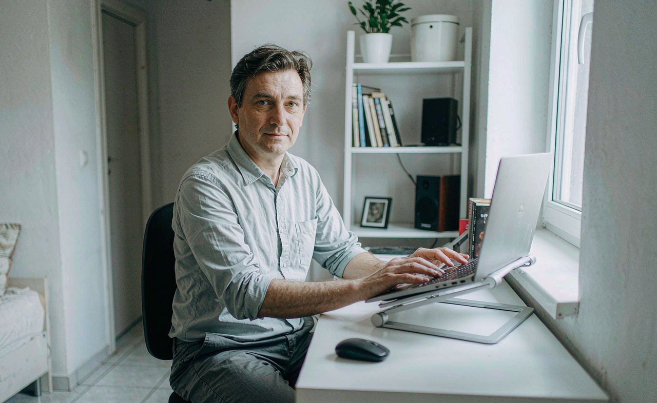 A man sitting upright at a tidy home office desk with a laptop, viewed from the side in soft daylight