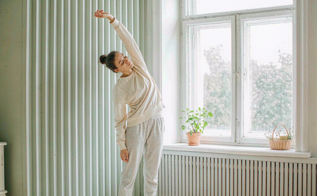 A woman gently stretching her neck and shoulders beside a bright living room window