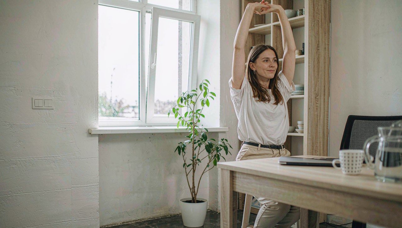 A person standing and stretching beside their desk in a bright home office with a laptop and notebook